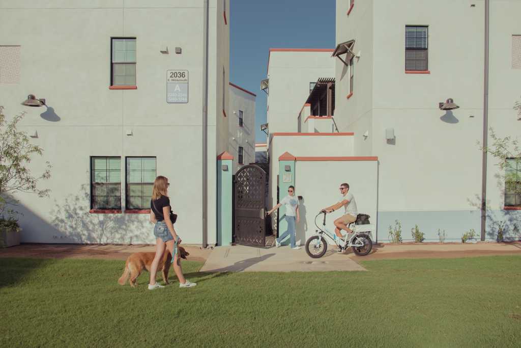 A car-free street at Culdesac Tempe: a woman walking a dog and a man on an e-bike between low white buildings